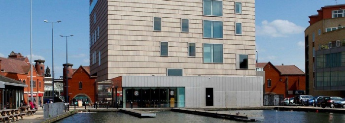 An image of the outside of Walsall Art Gallery. A tall grey building with water in the foreground and red brick buildings in the background.